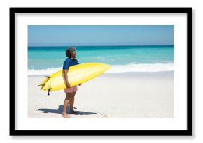Barefoot man is holding yellow surfboard and gazing toward turquoise ocean on white sand beach