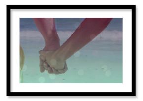 Holding hands of couple wearing light beach shorts, pink sleeveless top in shallow surf, soft flare