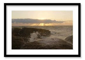 Crashing ocean waves spraying sea spray on jagged shore rocks, with sun rising behind low clouds