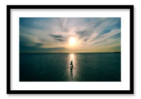 Little white boat floating on the water towards the horizon in the rays of the setting sun. Beautiful clouds with yellow highlights. Aerial view