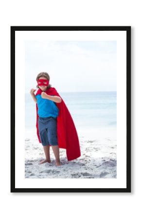 Child wearing red superhero cape and mask punching air on white sandy beach near calm sea