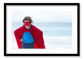 Child is standing on sandy beach wearing bright red cape and eye mask, ocean horizon visible