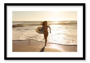 Woman running from breaking waves onto wet sand at beach edge, holding surfboard during golden hour