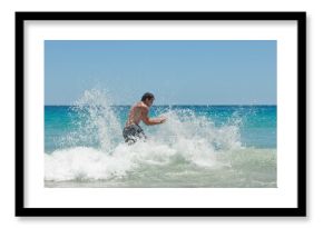 Man in dark swim shorts is wading in crashing turquoise waves at beach, sending water spray