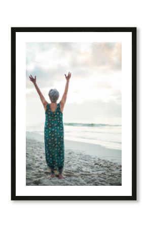 Barefoot senior woman raising arms toward sky on sandy beach by calm sea, under cloudy sky