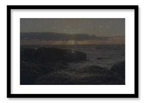 Sun sinking over horizon, casting glow on ocean waves crashing on rocky seashore under cloud layer
