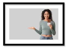 Happy young African-American woman with sweet chocolate bar showing something on grey background