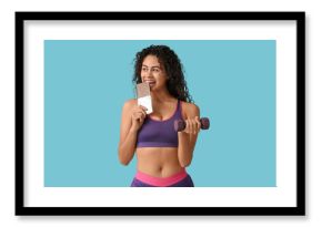 Beautiful young African-American woman with sweet chocolate bar and dumbbell on blue background