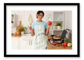 Beautiful young happy African-American woman with bell pepper and tongs cooking vegetables on modern electric grill in kitchen