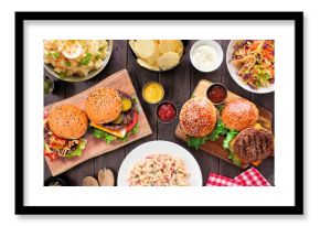 BBQ table scene with hamburgers, classic salads and snacks. Top view over a dark wood background.