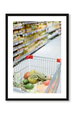 Shopping cart filled with fresh produce including pineapple, avocado, and oranges in supermarket aisle