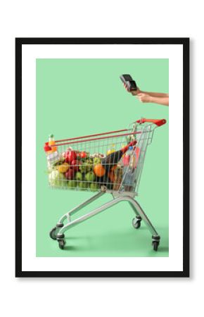 Female hand with payment terminal near shopping cart full of food on green background