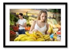 Young woman buyer choosing bunch of fresh bananas in vegetable shop