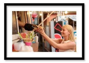 Joyful young blonde woman exchanges cash for food at lively outdoor market food truck with string stringlights, warm glow creating friendly, festive atmosphere
