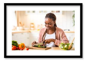 Happy young black woman cooking fresh vegetable salad in kitchen, chopping cucumber. Smiling millennial african american lady preparing lunch at home, enjoying eating healthy food, copy space