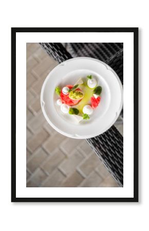 Gourmet appetizer with watermelon slice topped with avocado, herbs, and cream, elegantly plated on white dish in a stylish restaurant setting.