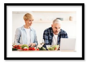Search for recipe on Internet, cook food and communicate with blog followers. Concentrated middle aged man work on laptop, woman preparing dish, on table with vegetables in modern kitchen interior