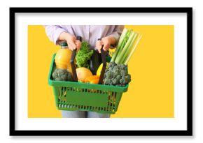 Woman holding green shopping basket with different fresh products on yellow background