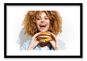 Joyful woman with curly hair holding a delicious burger, exuding happiness and enthusiasm against a plain background
