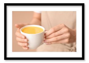 Young woman holding cup of tasty turmeric drink on beige background, closeup