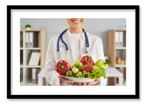 Nutritionist doctor holds vegetables promoting healthy diet. Smiling clinician with stethoscope offers a bowl of fresh salad in medical clinic. Promotes wellness healthcare and nutrition