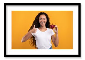 A happy millennial woman with curly hair stands against a yellow-orange background. She holds a red apple in one hand and gives a thumbs up with the other. She promotes healthy eating.