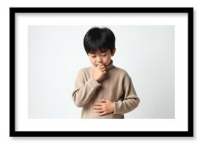 Young Asian boy wearing a beige turtleneck sweater is looking down with a pensive or slightly unwell expression. His hand is near his mouth and his other hand is on his stomach.
