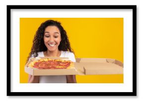 A joyful young lady gazes at an open pizza box filled with tasty Italian pizza. She stands in front of a bright yellow-orange wall, celebrating her food delivery.