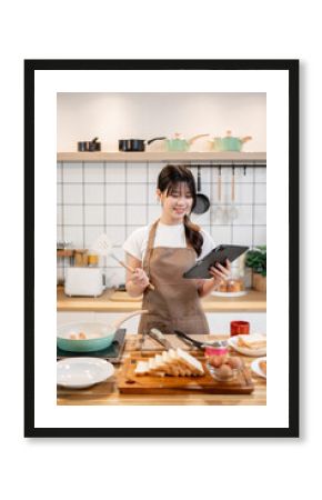 A beautiful Asian woman is looking at a recipe on her digital tablet while cooking in the kitchen.