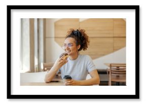Portrait of smiling young woman enjoying sweet doughnut and coffee in fast food restaurant.