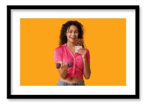 Happy young African-American woman with sweet chocolate bar and dumbbell on yellow background