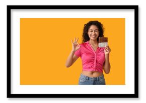 Happy young African-American woman with sweet chocolate bar showing ok gesture on yellow background