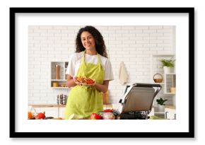 Beautiful young happy African-American woman holding plate with tasty vegetables near modern electric grill in kitchen