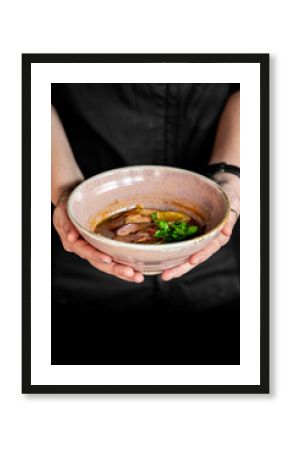 Close-up of hands holding a bowl of steaming soup with meat, vegetables, and herbs in a rustic ceramic dish, evoking warmth, comfort, and homemade food aesthetics.