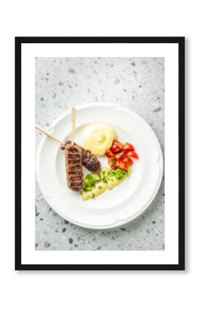 Grilled meat skewers with mashed potatoes, cherry tomatoes, and creamy herb sauce on a white plate over a gray speckled surface