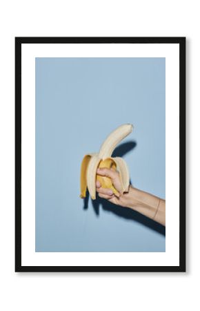 Caucasian young adult woman hand holding partially peeled banana against blue background, fingers gripping fruit firmly, nails painted, wrist adorned with thin bracelet, casting shadow