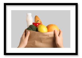 Female hands with vegetables, fruits, baguette and bottle of yogurt in paper bag on grey background