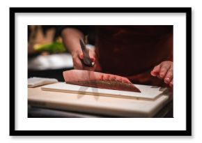 A chef's hands slice a thick tuna fillet with a yanagiba on a white board at a Japanese hotel sushi counter, dim focused lighting and warm tones emphasize craft.