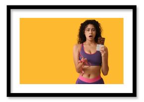 Shocked young African-American woman with sweet chocolate bar and measuring tape on yellow background