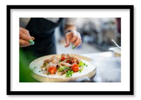 A close-up of a chef’s hands garnishing a gourmet dish with fresh green herbs. The plate features sliced meat, cherry tomatoes, and fresh greens, presented on a white plate