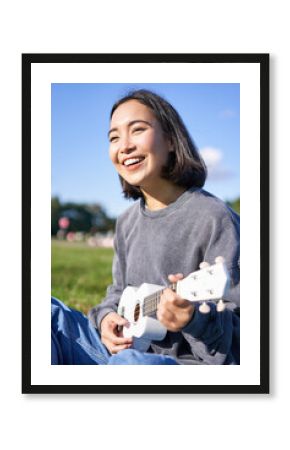 Vertical shot of smiling asian girl singing and laughing, playing ukulele, learn how to play instrument, sitting outdoors in park