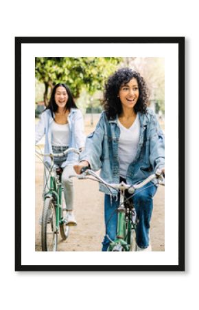 Vertical photo. Two young multiracial women having fun riding bike together in the park. Smiling female friends enjoying time together on summer vacation. Friendship and holidays concept.