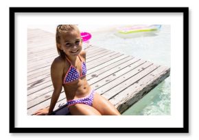 child wearing polka?dot bikini sitting on wooden dock, smiling beside pool float, copy space