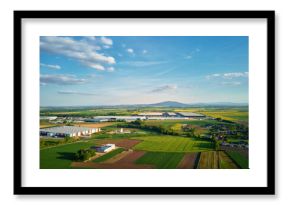 Aerial view of industrial warehouse complex near green agricultural fields and small village. Modern logistics infrastructure in rural landscape