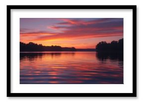 A peaceful lake at sunset featuring a glowing orange and purple sky. Dark clouds and distant mountains are mirrored on the water's surface, producing a tranquil and serene evening atmosphere.