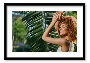 smiling woman with curly hair enjoying sunlight near tropical palm leaves, radiant skin and relaxed summer atmosphere in natural outdoor setting