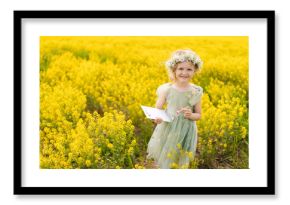 Cheerful child girl wearing flower wreath and fairy costume standing posing at blooming field during sunny summer day, drawing on canvas, enjoying beautiful nature. Concept of happiness childhood.