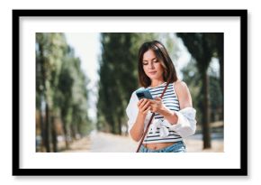 Young woman enjoying a sunny day at the park while texting on her phone, dressed in casual summer attire, carefree urban lifestyle