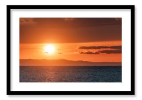 Golden sunset reflecting on calm sea with distant mountains and clouds creating a dramatic and peaceful atmosphere