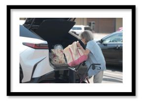 An elderly woman is grocery shopping while using a walker. She is loading her bags into the trunk of her car in a supermarket parking lot during the day.